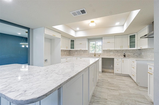 kitchen featuring visible vents, glass insert cabinets, wall chimney exhaust hood, tasteful backsplash, and a sink