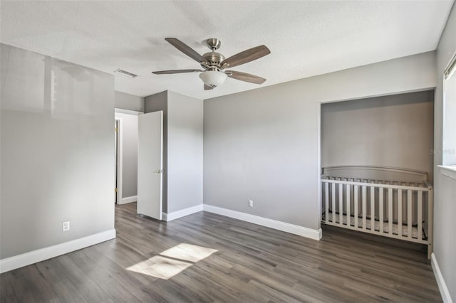 unfurnished bedroom featuring ceiling fan, a nursery area, dark hardwood / wood-style flooring, and a textured ceiling