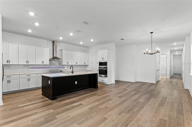 kitchen featuring a kitchen island with sink, wall chimney range hood, oven, light hardwood / wood-style floors, and white cabinetry