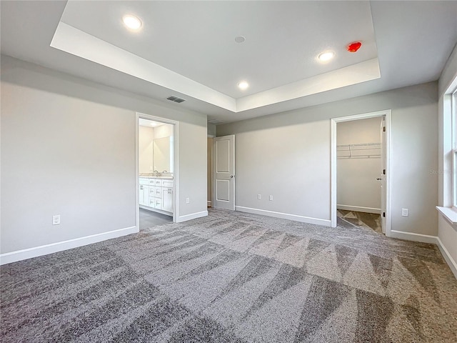 unfurnished bedroom featuring ensuite bath, a spacious closet, dark colored carpet, a raised ceiling, and a closet