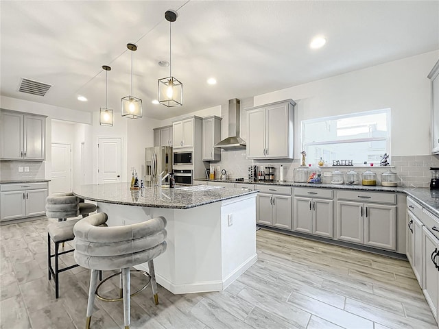 kitchen featuring a kitchen breakfast bar, stainless steel appliances, wall chimney range hood, pendant lighting, and an island with sink