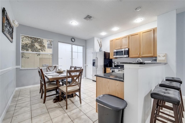 kitchen featuring kitchen peninsula, dark stone countertops, a textured ceiling, light brown cabinetry, and appliances with stainless steel finishes