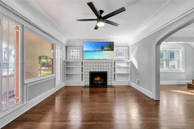 unfurnished living room with ceiling fan, a tiled fireplace, ornamental molding, built in shelves, and dark hardwood / wood-style flooring