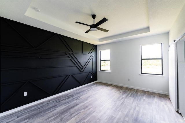 empty room with a raised ceiling, ceiling fan, a barn door, a textured ceiling, and light hardwood / wood-style floors