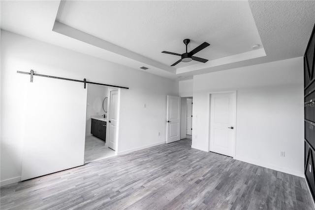unfurnished bedroom featuring ensuite bathroom, a tray ceiling, ceiling fan, a barn door, and light hardwood / wood-style floors