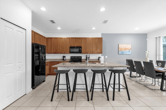 kitchen featuring a kitchen bar, black appliances, an island with sink, light tile patterned flooring, and light stone counters