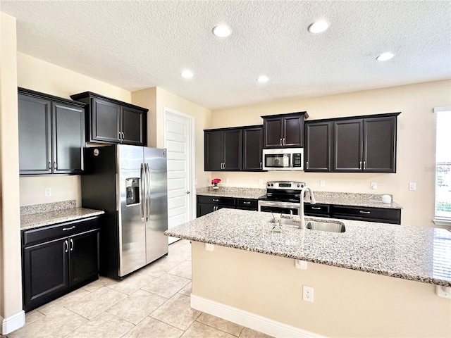 kitchen with a sink, a center island with sink, light stone counters, stainless steel appliances, and dark cabinets