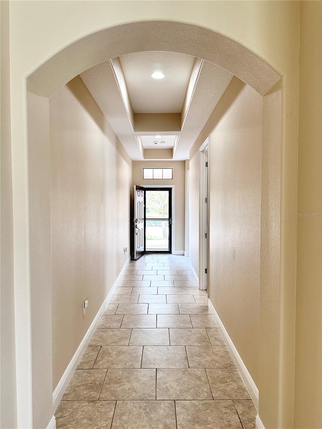 corridor featuring arched walkways, a tray ceiling, and baseboards