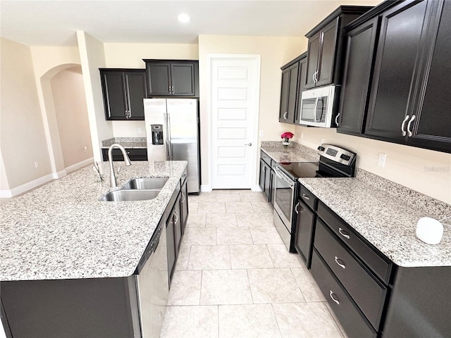 kitchen featuring arched walkways, a kitchen island with sink, a sink, appliances with stainless steel finishes, and dark cabinets