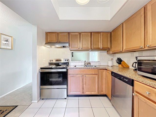 kitchen with a tray ceiling, appliances with stainless steel finishes, light tile patterned flooring, a sink, and under cabinet range hood