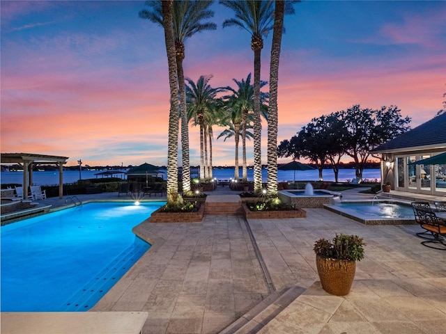 pool at dusk with a patio, pool water feature, a water view, and a hot tub