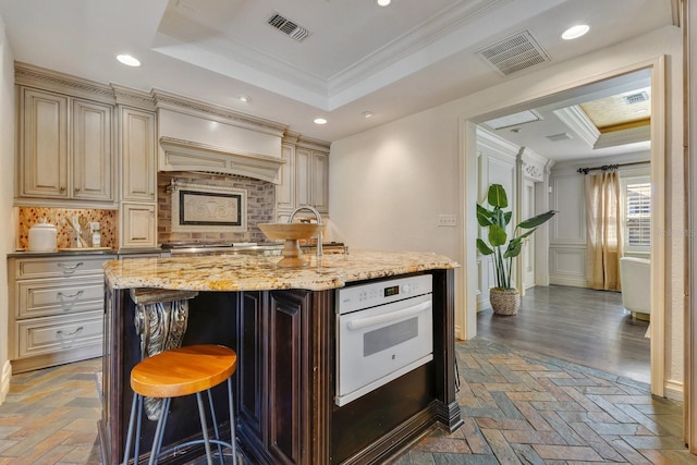 kitchen featuring backsplash, a tray ceiling, a kitchen island with sink, oven, and crown molding