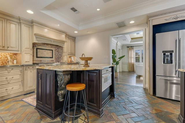 kitchen featuring a breakfast bar area, appliances with stainless steel finishes, a raised ceiling, a kitchen island with sink, and cream cabinets