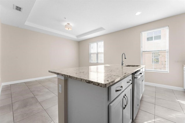 kitchen with sink, light stone counters, gray cabinets, a raised ceiling, and a kitchen island with sink