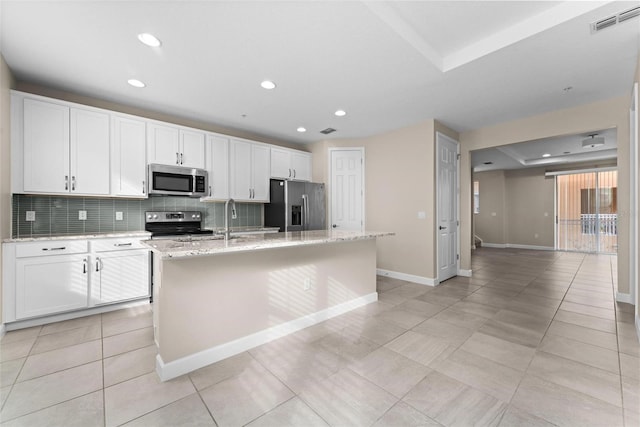 kitchen with appliances with stainless steel finishes, white cabinetry, backsplash, light stone counters, and a center island with sink