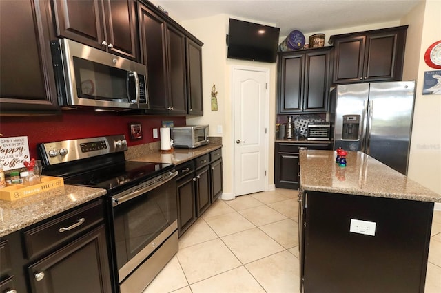 kitchen with light tile patterned floors, light stone counters, dark brown cabinets, a kitchen island, and appliances with stainless steel finishes