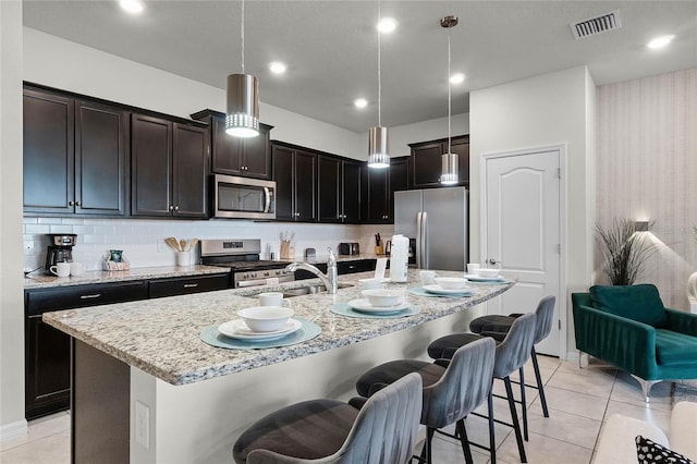 kitchen with appliances with stainless steel finishes, a center island with sink, light tile patterned floors, and decorative light fixtures