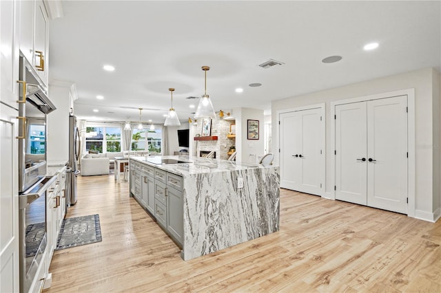 kitchen featuring white cabinetry, a large fireplace, hanging light fixtures, a large island with sink, and light stone counters