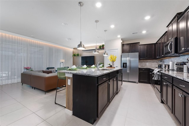 kitchen featuring decorative backsplash, light stone countertops, stainless steel appliances, a kitchen island with sink, and hanging light fixtures