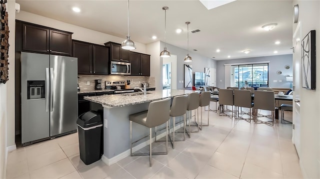 kitchen featuring decorative backsplash, light stone countertops, stainless steel appliances, a kitchen island with sink, and hanging light fixtures