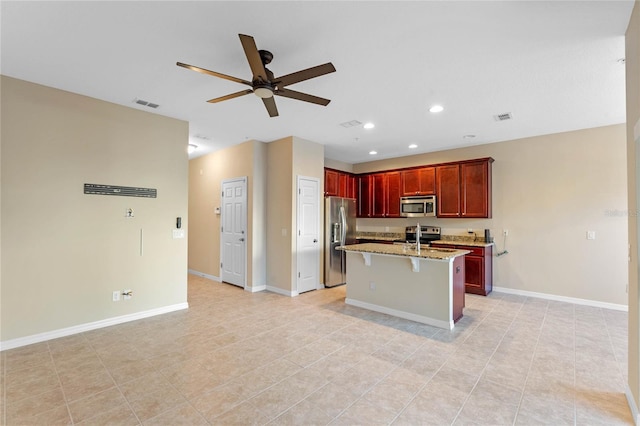 kitchen featuring ceiling fan, light stone countertops, an island with sink, appliances with stainless steel finishes, and a breakfast bar area