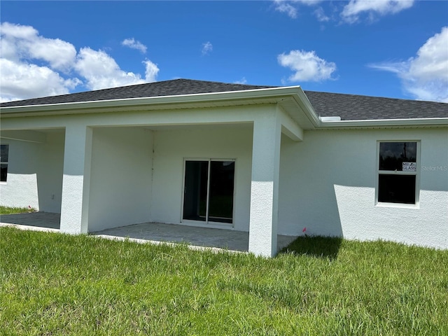 rear view of property featuring a patio, a lawn, roof with shingles, and stucco siding