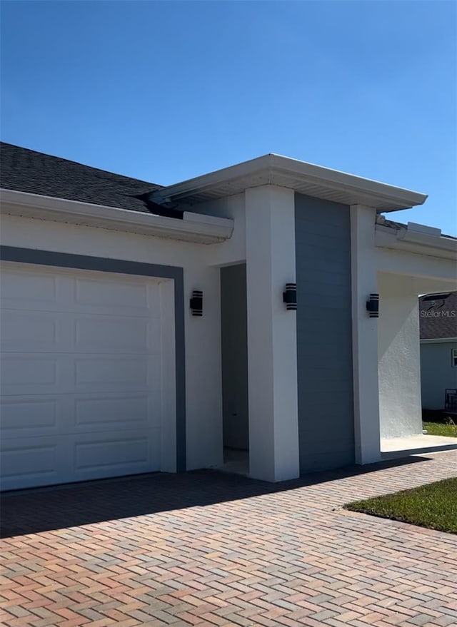 exterior space featuring decorative driveway, an attached garage, roof with shingles, and stucco siding