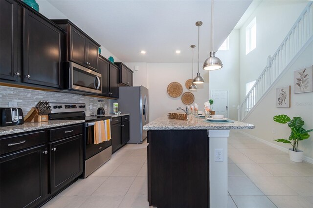 kitchen with hanging light fixtures, stainless steel appliances, tasteful backsplash, light stone counters, and a kitchen island with sink