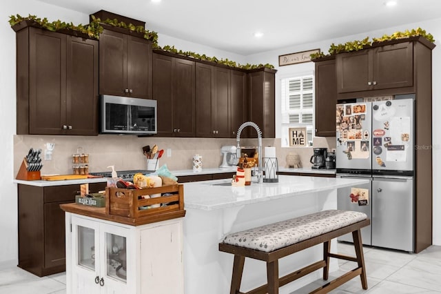 kitchen with decorative backsplash, dark brown cabinetry, stainless steel appliances, and a center island with sink