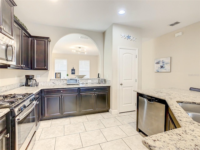 5303 Wildwood Way, Davenport, FL 33837 - kitchen with stainless steel appliances, arched walkways, light stone counters, dark brown cabinetry, and light tile patterned floors