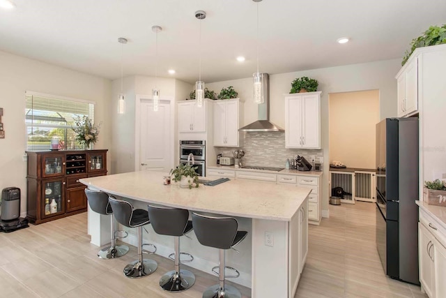 kitchen featuring white cabinetry, wall chimney exhaust hood, refrigerator, decorative light fixtures, and a kitchen island with sink