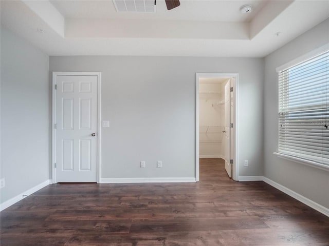 unfurnished bedroom featuring dark wood-type flooring, a walk in closet, a tray ceiling, a closet, and ceiling fan