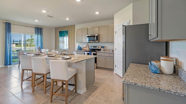 kitchen with light stone counters, an island with sink, light tile patterned floors, and appliances with stainless steel finishes