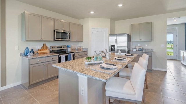 kitchen with stainless steel appliances, light stone counters, a kitchen bar, a kitchen island with sink, and light tile patterned floors