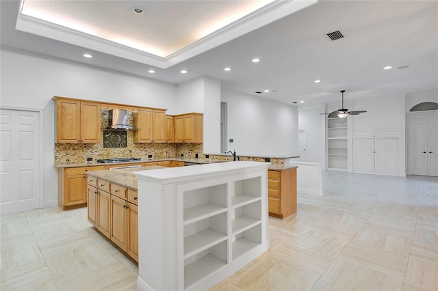 kitchen featuring ceiling fan, wall chimney range hood, kitchen peninsula, stainless steel gas stovetop, and a kitchen island