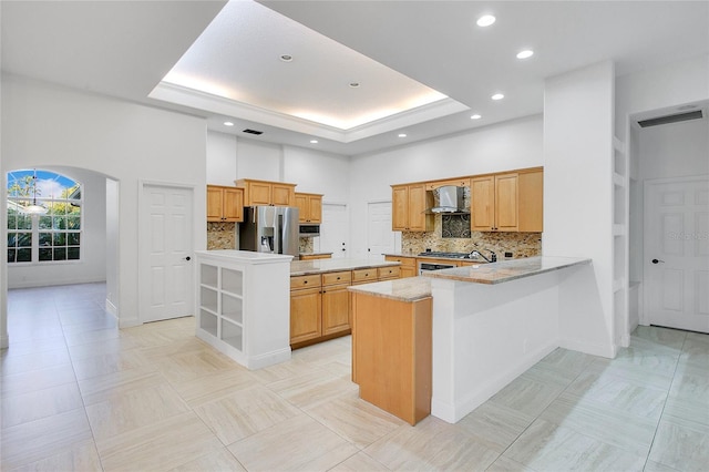 kitchen with kitchen peninsula, stainless steel refrigerator with ice dispenser, a towering ceiling, backsplash, and wall chimney range hood