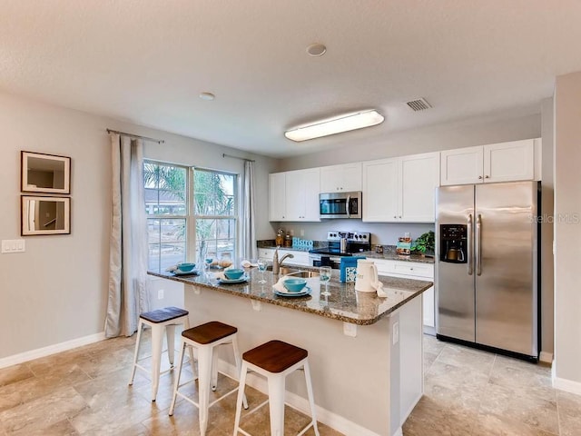 kitchen with a kitchen bar, appliances with stainless steel finishes, dark stone countertops, white cabinets, and an island with sink