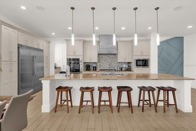 kitchen featuring a spacious island, wall chimney range hood, built in appliances, light stone countertops, and decorative light fixtures