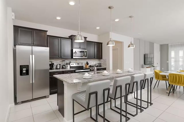 kitchen with light stone countertops, hanging light fixtures, stainless steel appliances, a kitchen bar, and a kitchen island with sink