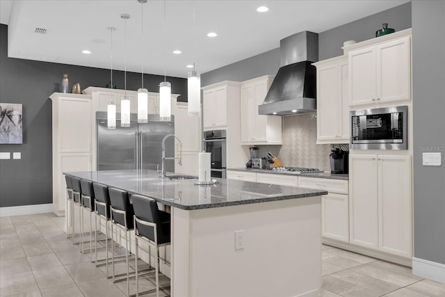 kitchen featuring wall chimney range hood, sink, hanging light fixtures, built in appliances, and an island with sink