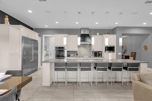 kitchen with wall chimney range hood, built in appliances, decorative light fixtures, white cabinetry, and an island with sink