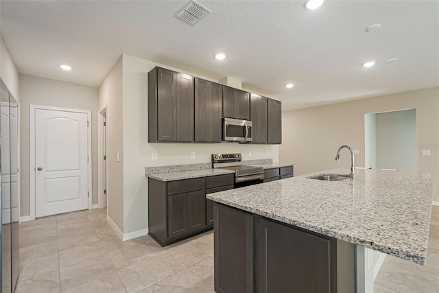 kitchen with a kitchen island with sink, sink, light stone countertops, appliances with stainless steel finishes, and dark brown cabinetry