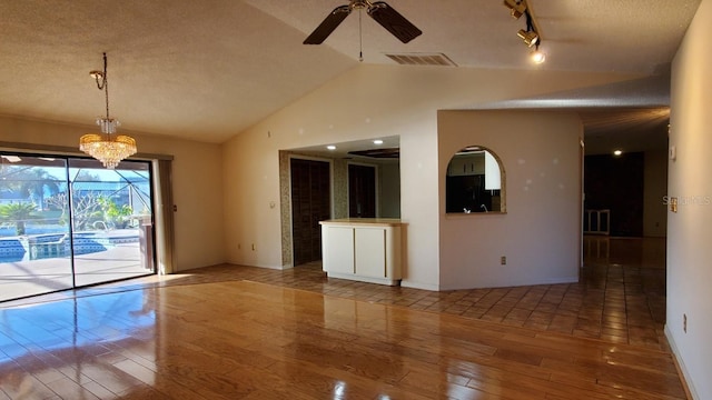 empty room with ceiling fan with notable chandelier, rail lighting, vaulted ceiling, hardwood / wood-style flooring, and a textured ceiling