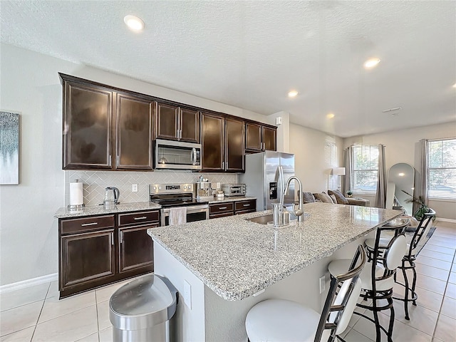 kitchen with appliances with stainless steel finishes, dark brown cabinets, sink, light tile patterned floors, and an island with sink