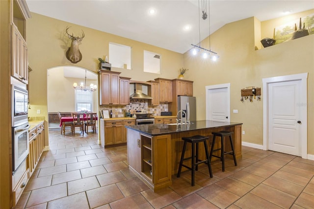 kitchen with high vaulted ceiling, hanging light fixtures, wall chimney exhaust hood, an island with sink, and appliances with stainless steel finishes