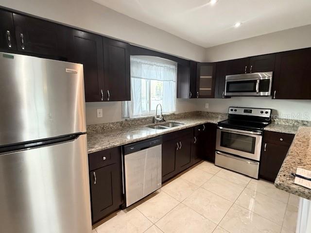 kitchen with dark brown cabinetry, light stone countertops, sink, light tile patterned flooring, and appliances with stainless steel finishes