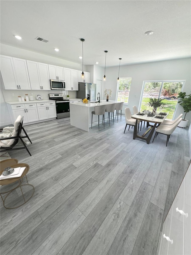 kitchen featuring light wood-type flooring, an island with sink, appliances with stainless steel finishes, decorative light fixtures, and white cabinetry