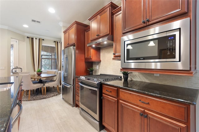 kitchen with under cabinet range hood, visible vents, appliances with stainless steel finishes, ornamental molding, and dark stone counters