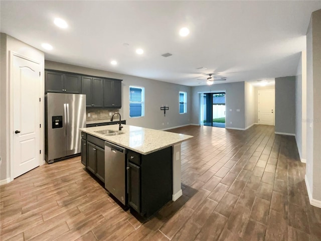 kitchen with ceiling fan, sink, light stone counters, a center island with sink, and appliances with stainless steel finishes