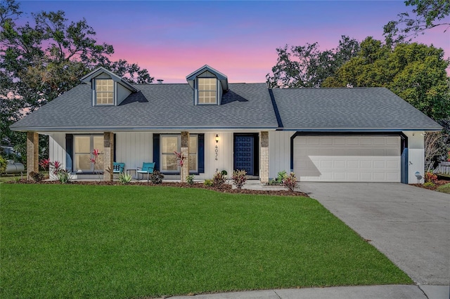 new england style home with a porch, a yard, concrete driveway, a shingled roof, and a garage